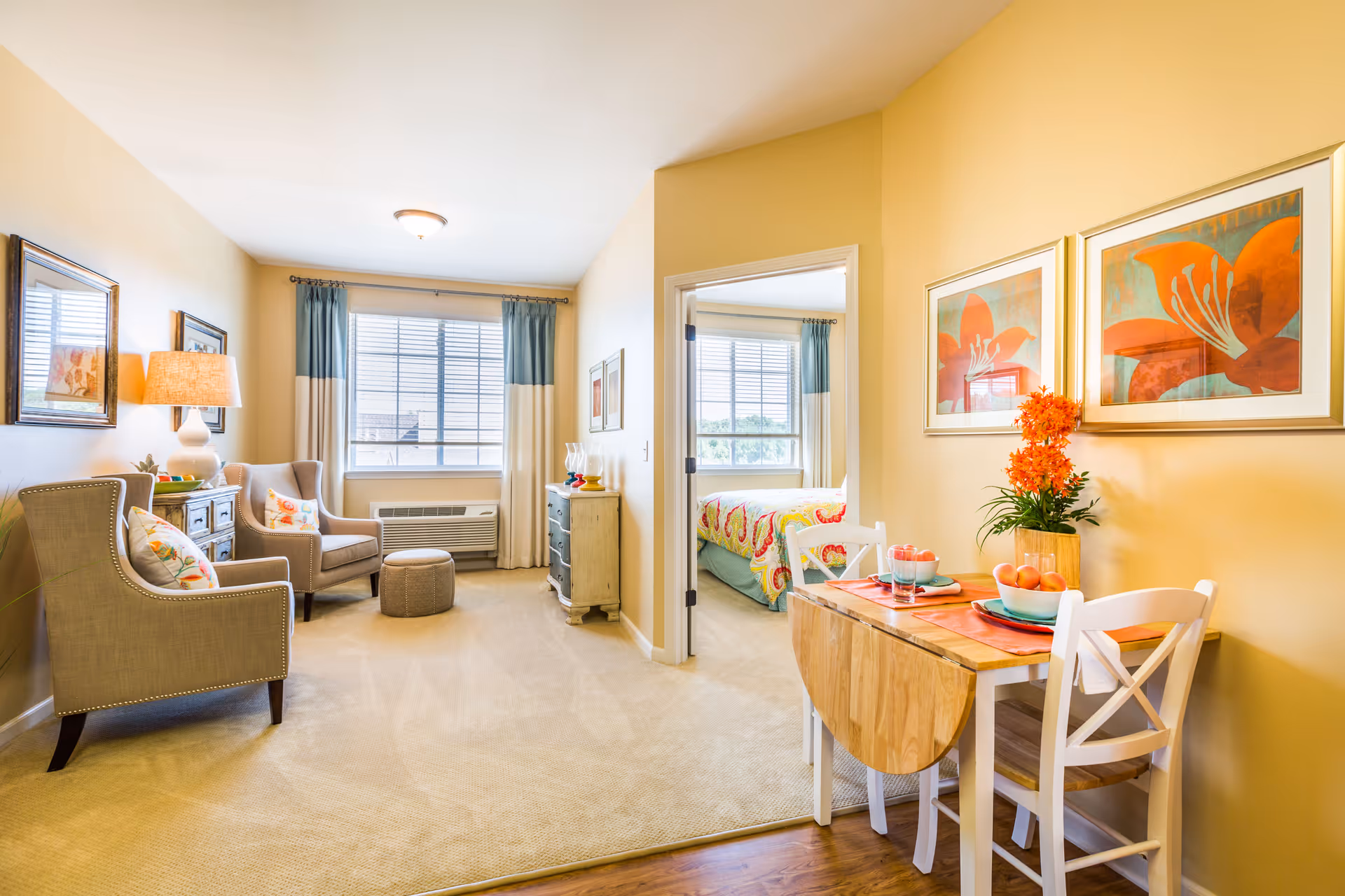 A bright and cozy senior living apartment interior featuring a small dining area with a wooden table set for two, decorated with orange flowers and fruit. Adjacent to the dining area is a living room with two upholstered armchairs, a small ottoman, a side table with a lamp, and framed artwork on the walls. In the background, a bedroom with a colorful bedspread is visible through an open doorway. Large windows with curtains allow natural light to fill the space.