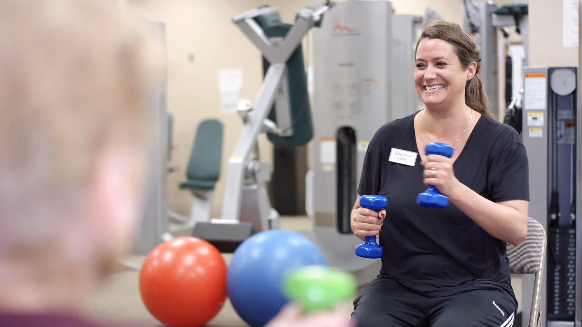 A smiling woman holding blue dumbbells is seated in a fitness room with exercise equipment and colorful exercise balls in the background. Another person is partially visible in the foreground.