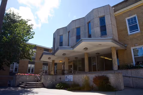 Exterior view of the Prospect Heights Community Living Center building entrance with a covered porch supported by columns, surrounded by windows and landscaping including bushes and flowers, under a partly cloudy sky.
