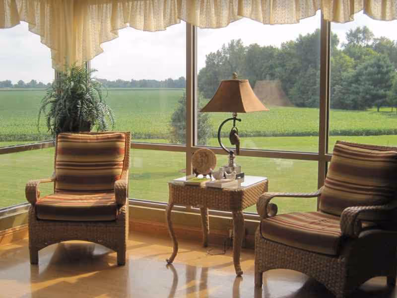A cozy seating area with two cushioned wicker chairs and a small wicker side table between them, holding a decorative lamp and small items. The area is next to large windows with a view of green fields and trees outside, with a light-colored valance curtain at the top.
