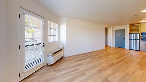 Empty interior room with light wood flooring, white walls, a door with window blinds leading outside, a window with blinds, a wall-mounted heater, and a small kitchen area with a refrigerator and cabinets in the background.