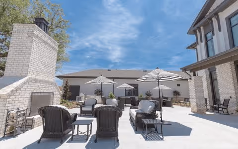 Outdoor patio area with multiple seating arrangements including cushioned chairs and small tables. There are several striped umbrellas providing shade. A large white brick fireplace is visible on the left side. The sky is clear and blue, and the building walls are light-colored with windows and doors.