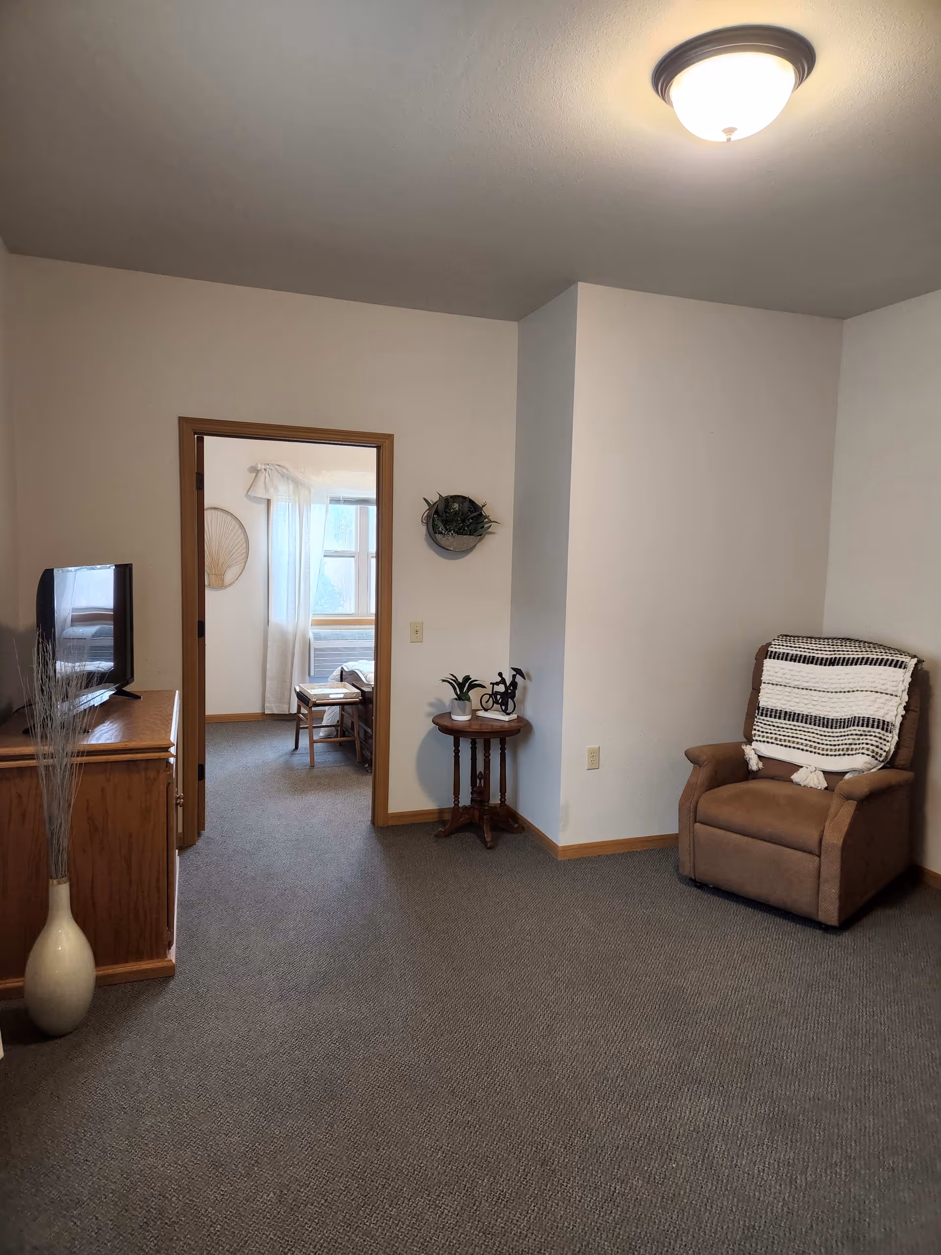 A cozy living room area with a brown recliner chair draped with a black and white throw blanket, a small round wooden side table with two small plants and a decorative bicycle, and a wooden cabinet with a flat-screen TV on top. The room has beige walls, a ceiling light fixture, and carpeted floor. Through an open doorway, a bright room with a window, white curtains, and a wooden chair is visible.