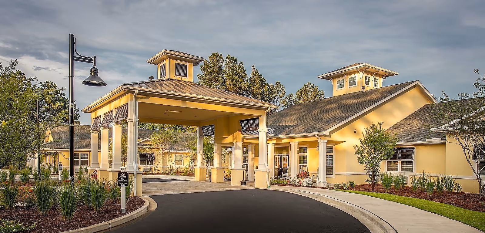 Exterior view of The Addison of West Ashley facility showing a covered entrance with columns, a curved driveway, landscaped greenery, and a yellow building with multiple windows under a partly cloudy sky.