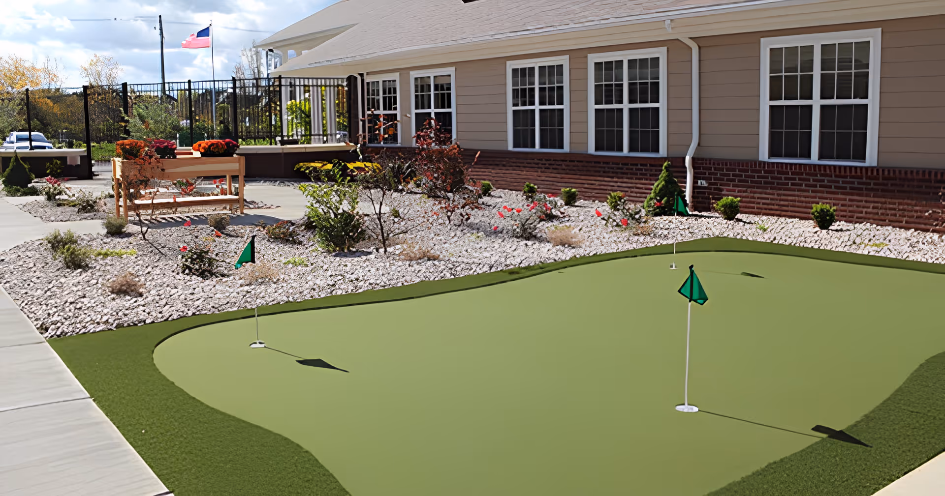 Outdoor putting green with three small flags in front of a beige building with white-trimmed windows. There are landscaped areas with rocks, bushes, and flowers surrounding the putting green, and a wooden bench with flower pots is visible near the building. A flagpole with an American flag is in the background.