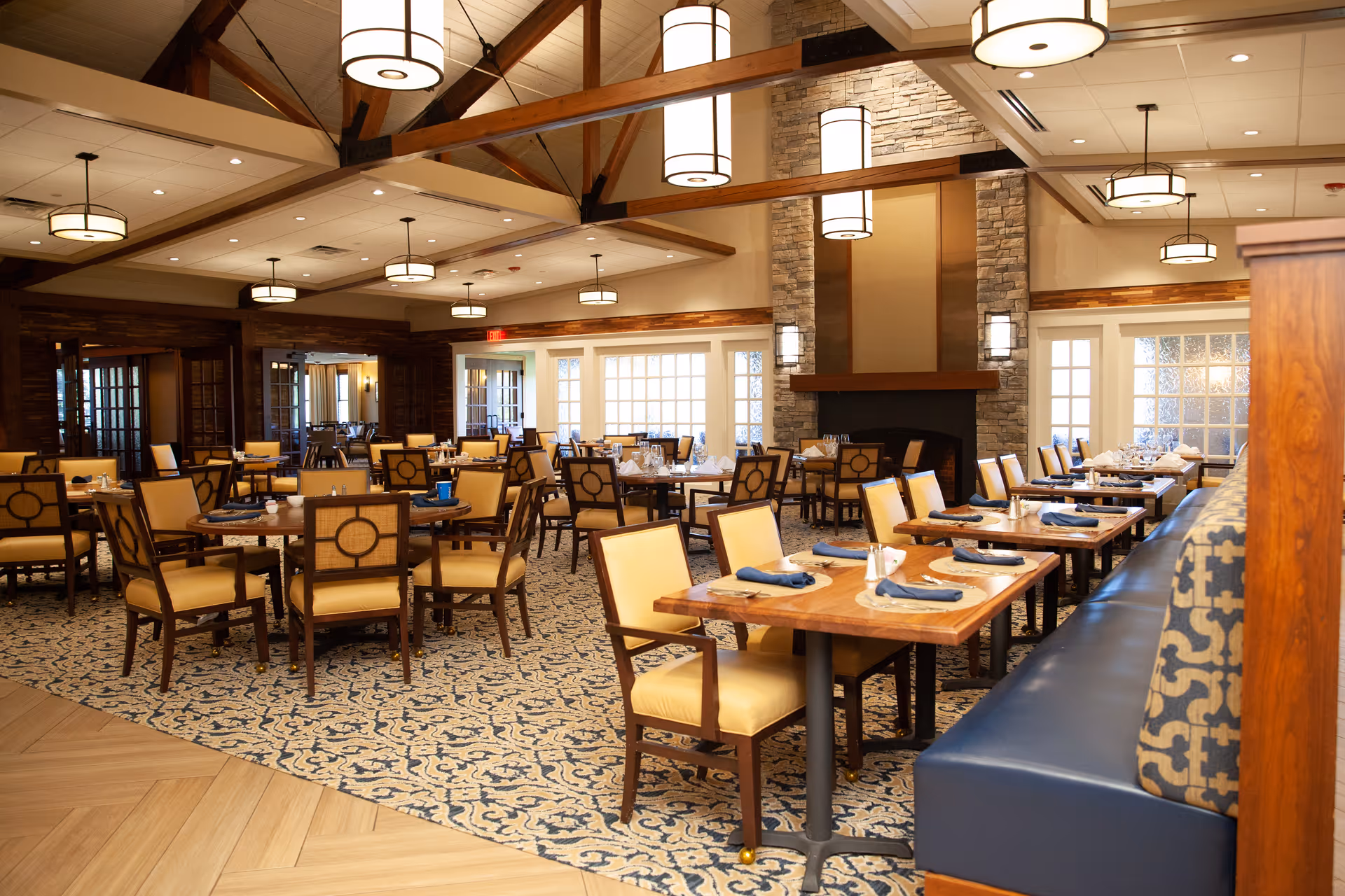 Spacious, well-lit dining room featuring wooden tables and upholstered chairs, booth seating, patterned carpet, and a stone fireplace.