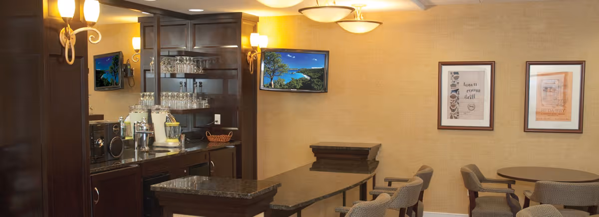Interior view of a lounge area with a dark wood bar counter, glass shelves with glassware, a microwave, and a blender. There are several chairs around a small round table and a long counter with stools. The walls are beige with framed artwork and a flat-screen TV mounted on one wall. Warm lighting fixtures are mounted on the walls.