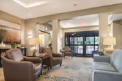 A cozy and well-lit seating area in a senior living facility featuring leather armchairs and a sofa arranged around a patterned rug. The space includes side tables with lamps, decorative artwork on the walls, and large glass doors leading outside.