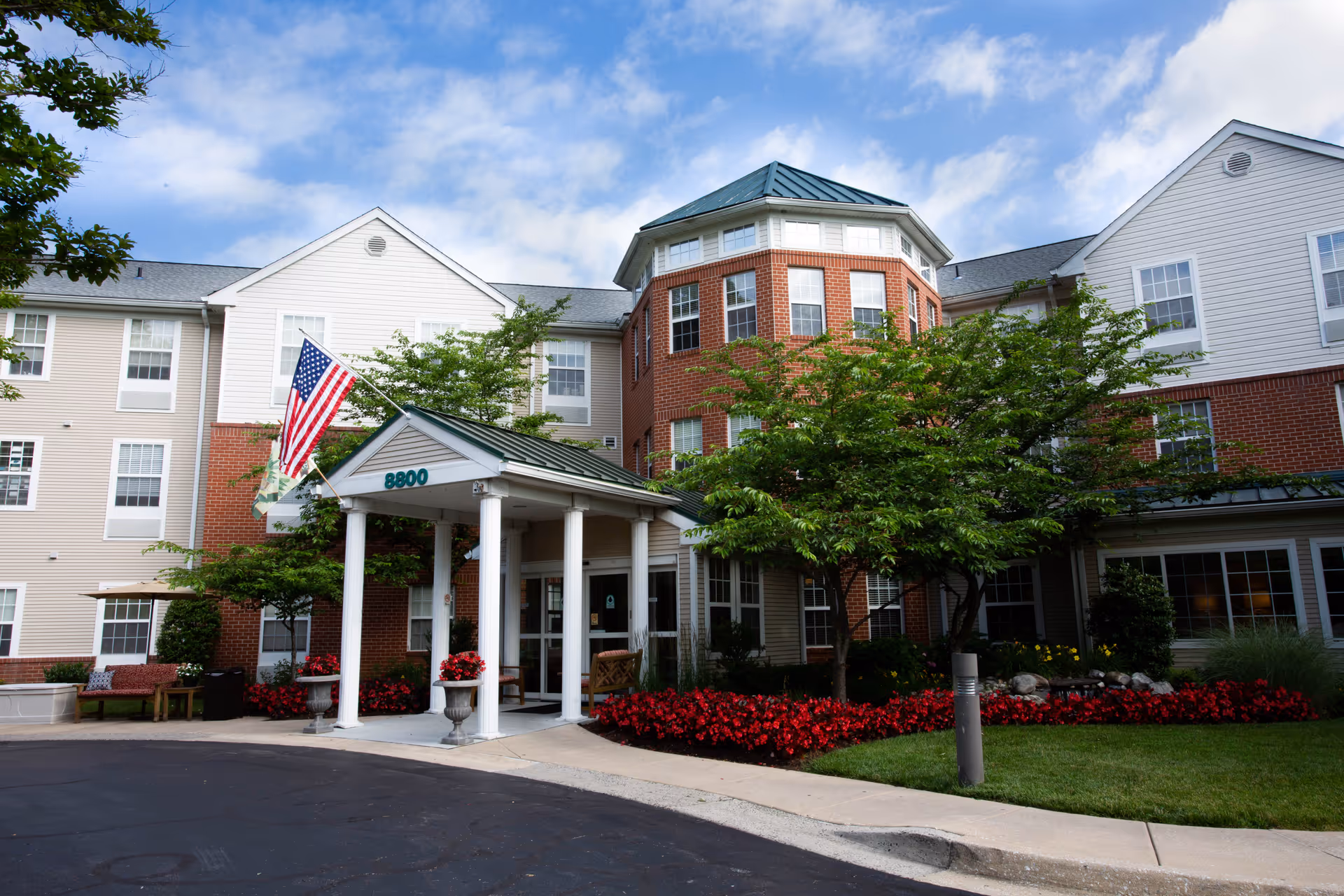 Exterior view of Morningside House of Satyr Hill, a multi-story senior living facility with a brick and white siding facade, a covered entrance with white columns, American flag, well-maintained landscaping including trees and red flowers, and a clear blue sky with some clouds.