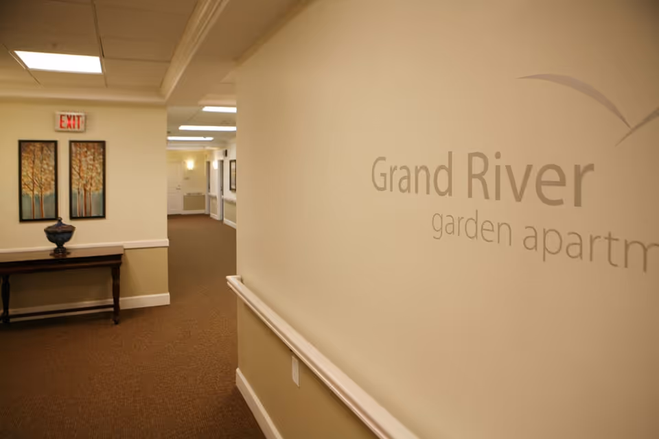 Carpeted interior hallway of a senior living facility with 'Grand River garden apartments' wall lettering, a console table with a vase, and an EXIT sign.