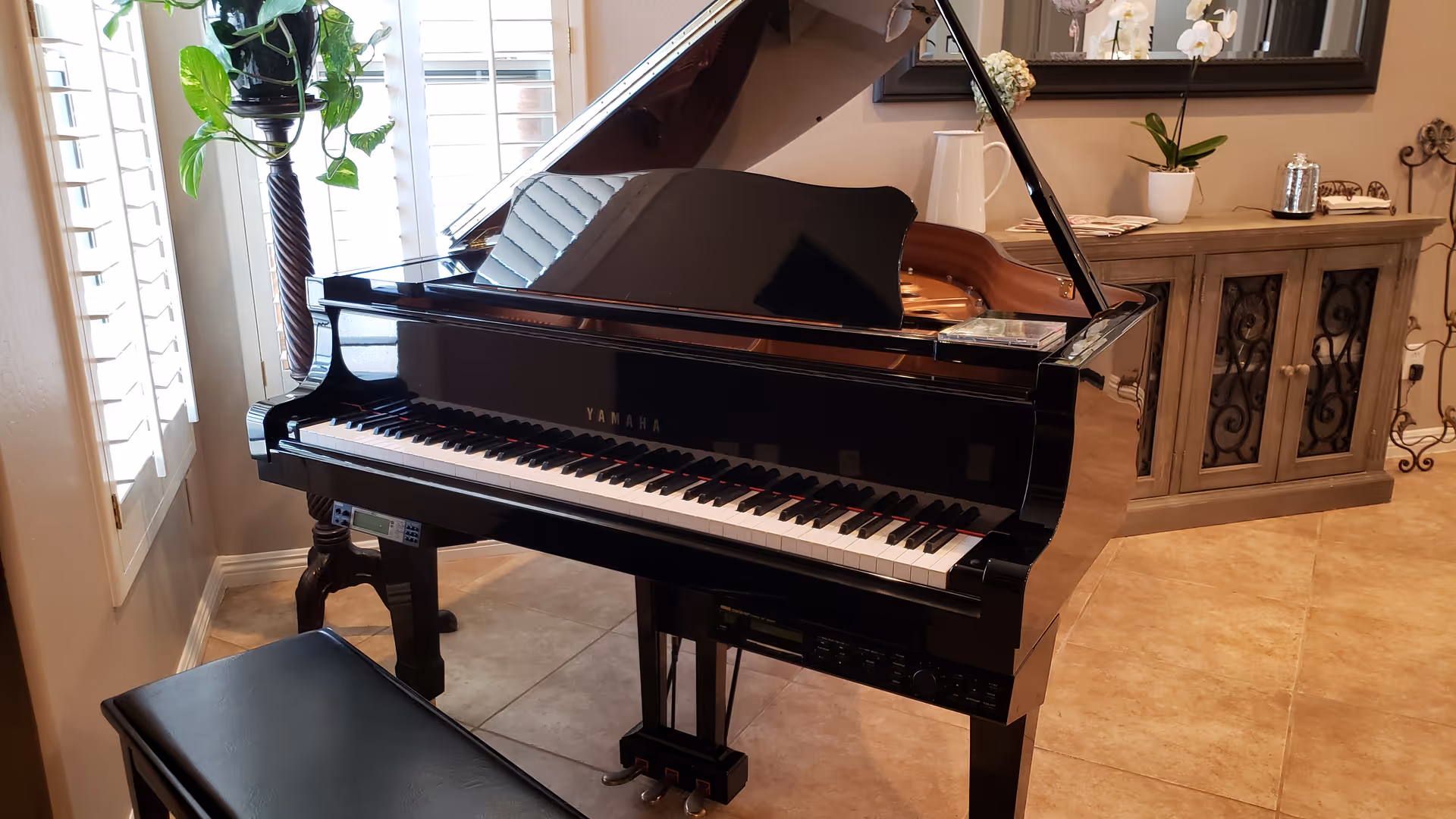 A black Yamaha grand piano with its lid open, placed in a room with beige tiled flooring. To the left, there is a tall plant stand with a green leafy plant near white window shutters. In the background, there is a wooden cabinet with decorative items including a white orchid and a large mirror above it.