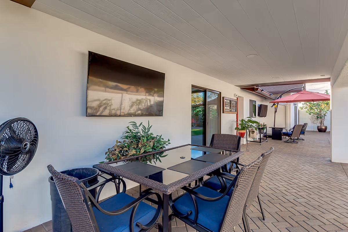 Covered outdoor patio area with a glass-top table and four wicker chairs with blue cushions. A wall-mounted flat screen TV is visible on the white wall. There are potted plants, a standing fan, and a red umbrella shading additional seating further down the patio. The floor is paved with brick tiles.