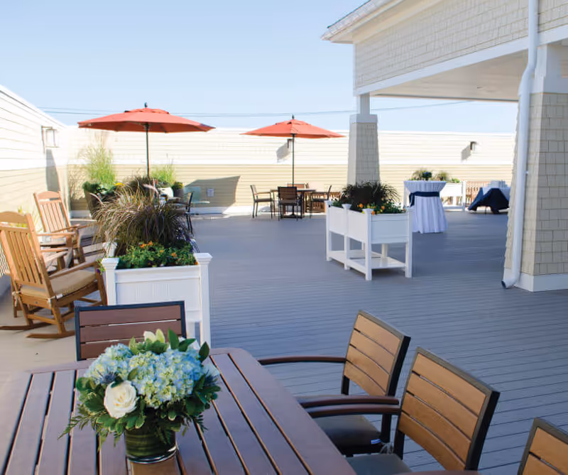 Outdoor patio area with wooden tables and chairs, potted plants, and red umbrellas providing shade. The space is spacious with a light gray deck floor and beige walls, featuring a covered section with columns and additional seating.
