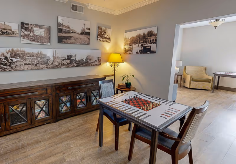 A cozy interior room with a wooden table featuring a chess and backgammon board, surrounded by two chairs. Against the wall is a dark wooden cabinet with glass doors displaying books or magazines. Above the cabinet are several black and white vintage photographs. A floor lamp with a yellow shade is turned on, casting warm light. In the adjacent room, there is a beige armchair and a table with a lamp, all set on light wood flooring.