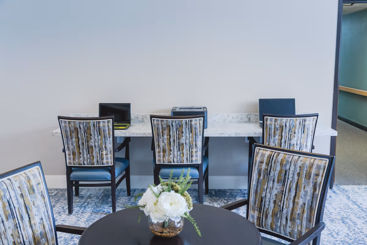 A small computer workstation area in a senior living facility with three chairs facing a marble countertop holding two laptops and a printer. In the foreground, there is a round table with a floral arrangement and two patterned chairs. The walls are light-colored and the floor has a blue patterned carpet.