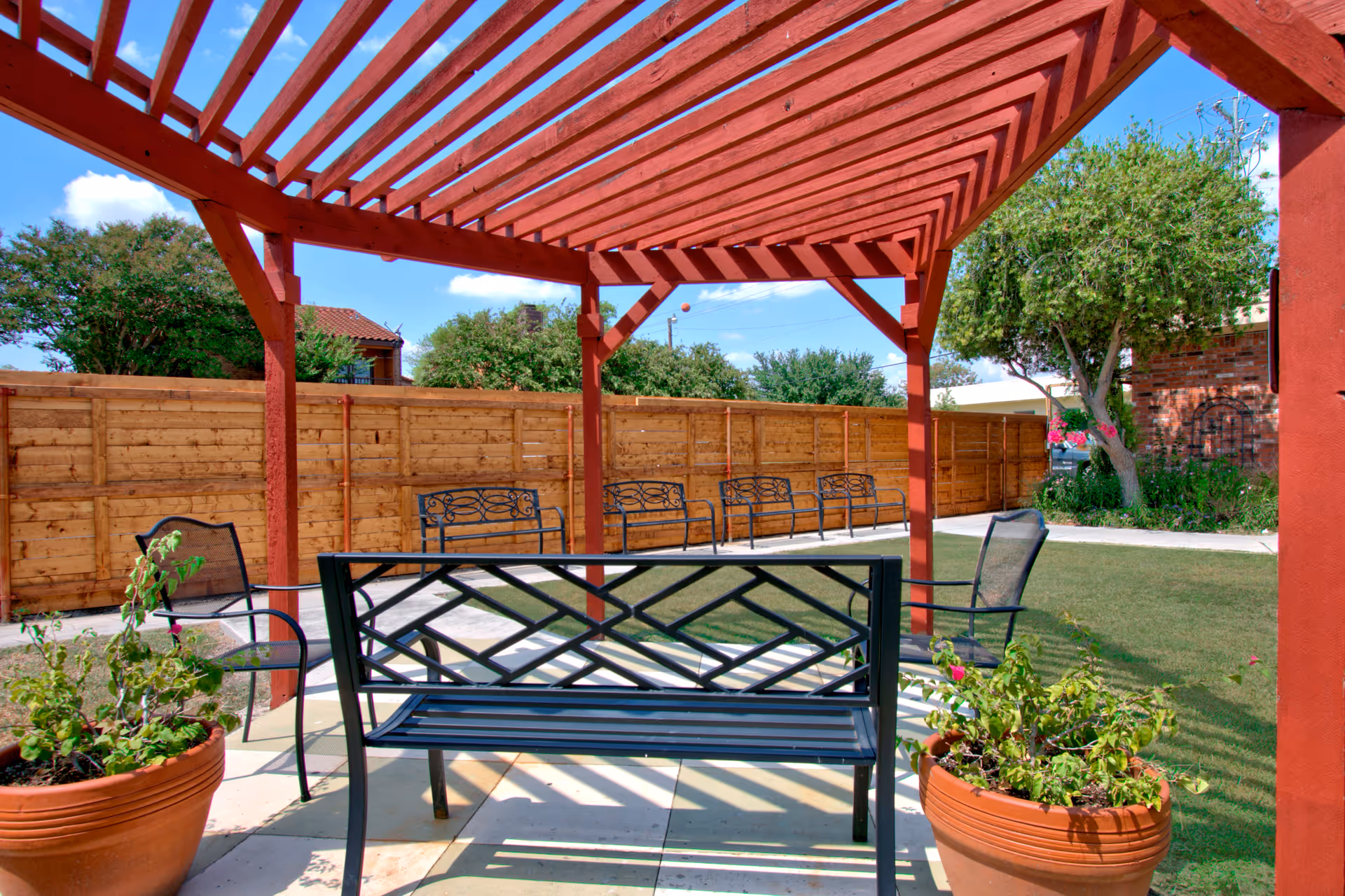 Outdoor seating area with a red wooden pergola providing partial shade. There are black metal benches and chairs arranged on a paved surface, with two large terracotta pots containing plants in the foreground. A wooden fence and green trees are visible in the background under a blue sky with some clouds.