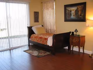 Bedroom in a senior facility with dark wood bed, nightstand, lamp, and sliding glass doors.