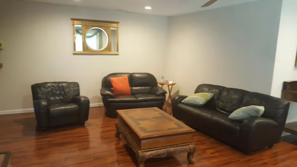 A living room with dark leather furniture including a single armchair, a loveseat with an orange pillow, and a sofa with two light green pillows. There is a wooden coffee table with ornate legs in the center on a polished wooden floor. A gold-framed mirror with a round center hangs on the light-colored wall behind the loveseat. A small wooden side table with a water bottle and a few items is next to the loveseat.