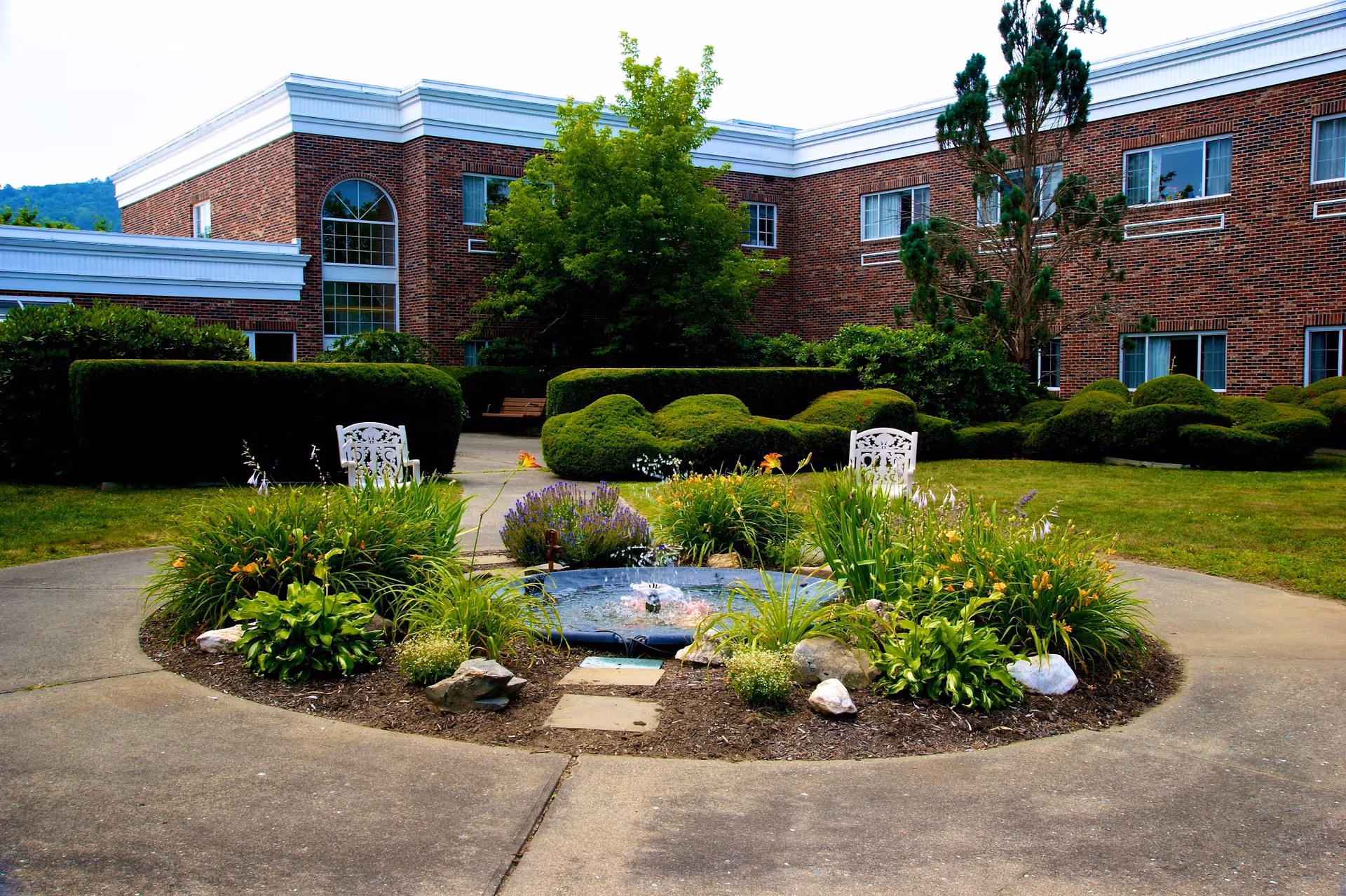 A landscaped courtyard with a fountain surrounded by bushes and flowers in front of a brick building.