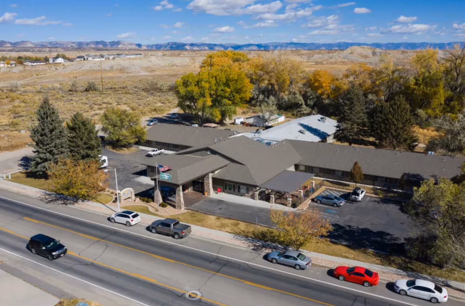 Aerial view of Pinnacle Nursing and Rehabilitation Center, a single-story building with a large parking lot and several cars parked along the street in front. The facility is surrounded by trees with autumn foliage and open land in the background under a partly cloudy sky.