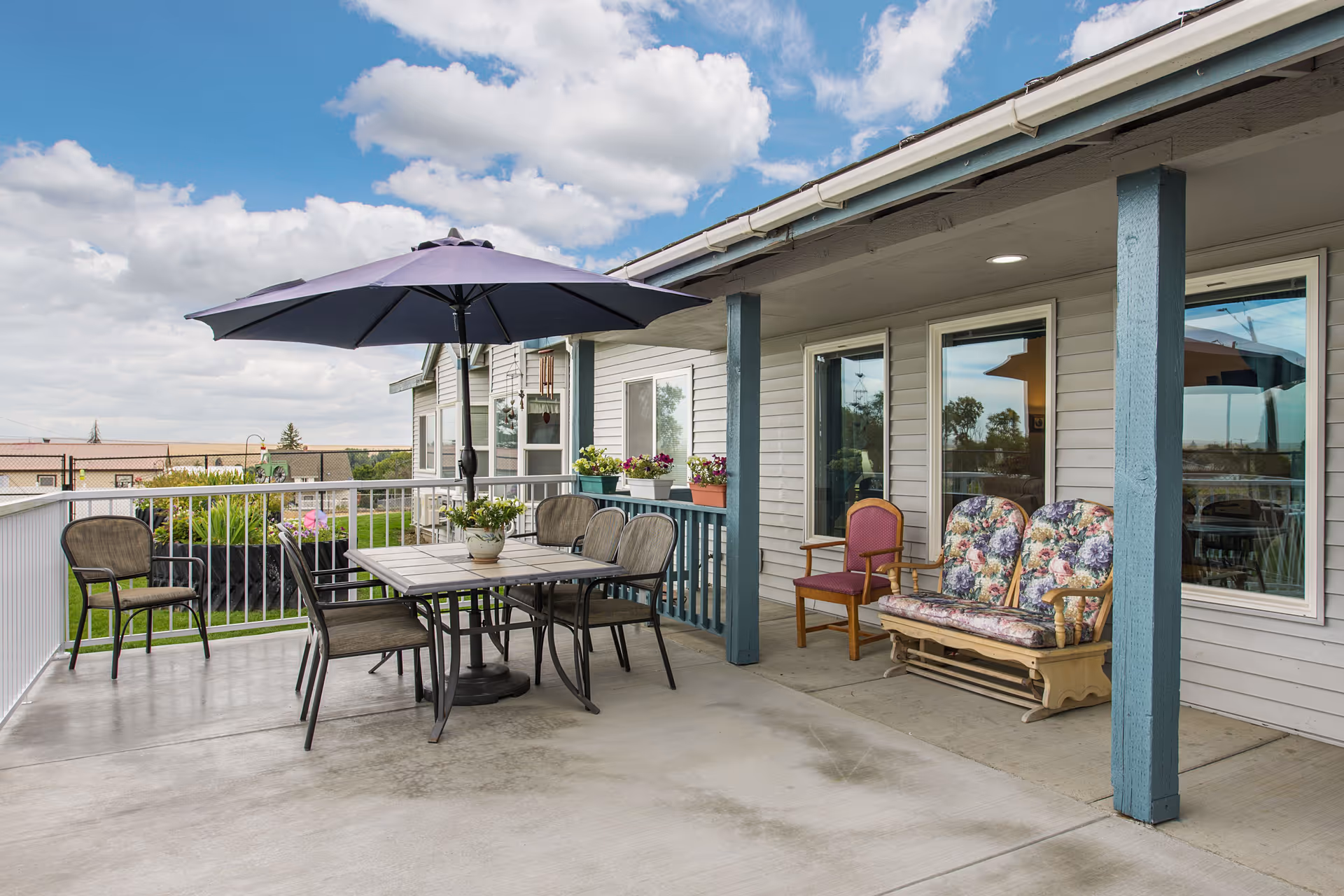 Outdoor patio area at Vista Manor with a table and six chairs under a large purple umbrella. There are additional chairs including a floral cushioned bench and a single chair with a red cushion. The patio is adjacent to the building with windows and flower pots on the railing. The sky is partly cloudy.