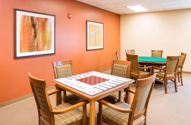 A room with two wooden game tables and several wooden chairs with striped cushions. One table has a checkerboard and backgammon design, and the other table has a green felt surface. The walls are painted orange and beige, with two framed abstract paintings hanging on the orange wall. The room has a beige carpet and a white ceiling with recessed lighting.