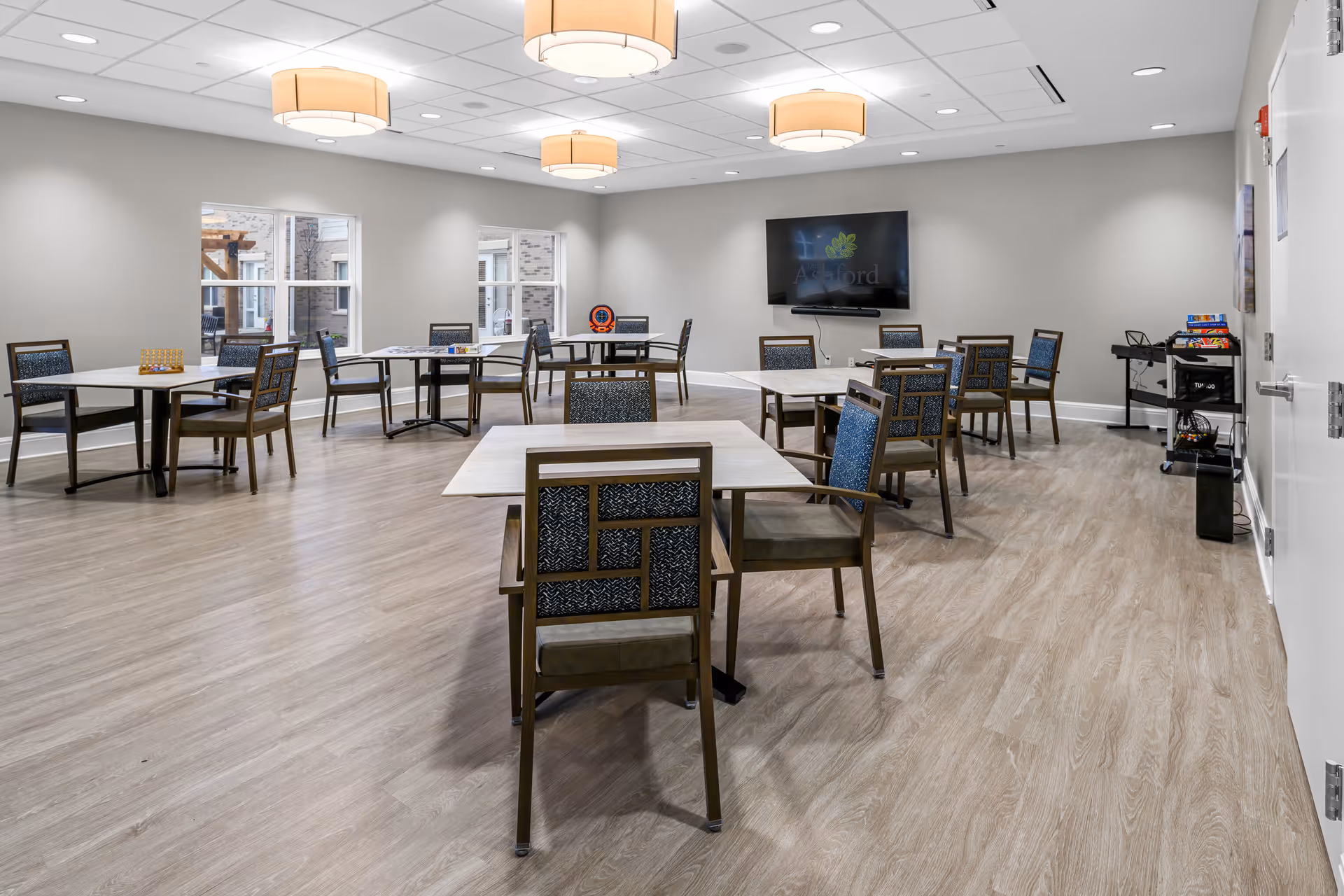 A spacious, well-lit common room with multiple square tables and chairs arranged for group seating. The room has light wood flooring, neutral-colored walls, and large windows letting in natural light. Ceiling lights provide additional illumination. A large flat-screen TV is mounted on the far wall displaying the Ashford logo. A cart with board games and other recreational items is visible near the door.