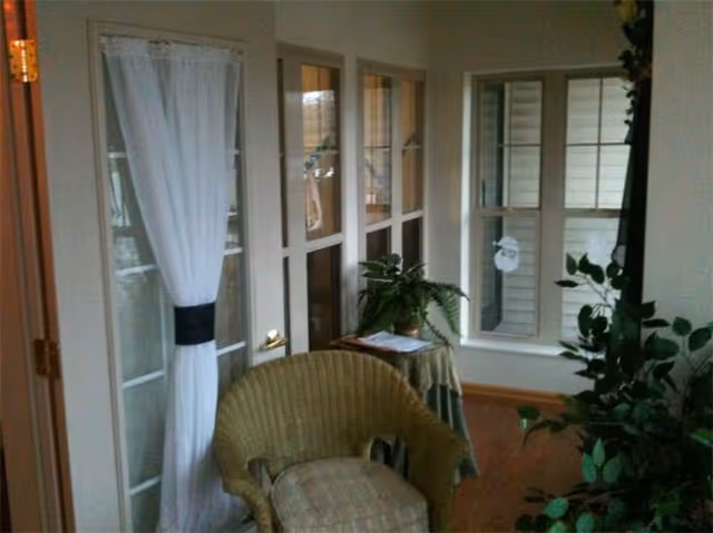 A cozy indoor seating area with a wicker chair in the foreground, a small table covered with a cloth and a potted plant on top, and multiple windows with white curtains letting in natural light.