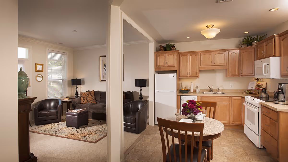 Interior view of a senior living facility showing a small kitchen and adjacent living room. The kitchen has wooden cabinets, a white refrigerator, stove, microwave, and a round dining table with two chairs and a flower centerpiece. The living room has a dark sofa, two leather armchairs, a patterned rug, a side table with a lamp, and a window with blinds.