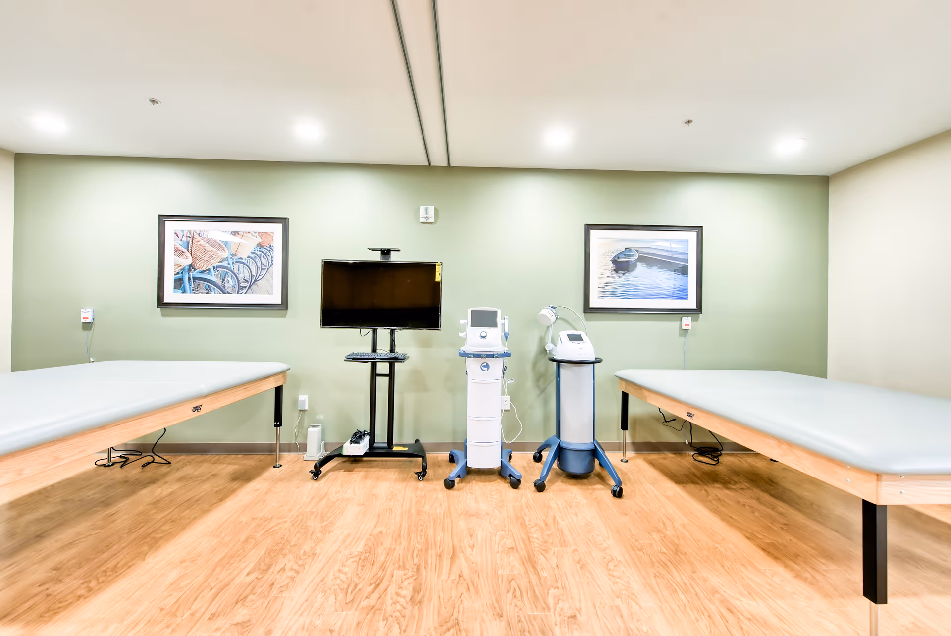 A medical treatment room with two padded examination tables on either side, a monitor on a stand, and two medical devices on wheeled carts against a green wall. The room has wooden flooring and two framed pictures hanging on the wall above the equipment.
