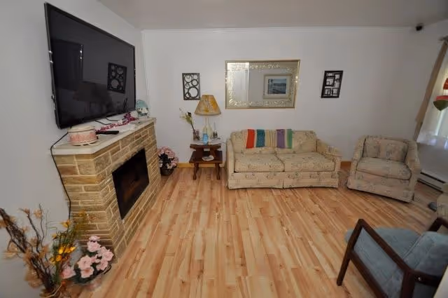 A cozy living room with light wood flooring, a stone fireplace with a mounted flat-screen TV above it, a beige sofa with a colorful throw blanket, a matching armchair, a wooden side table with a lamp, and framed artwork on the walls.