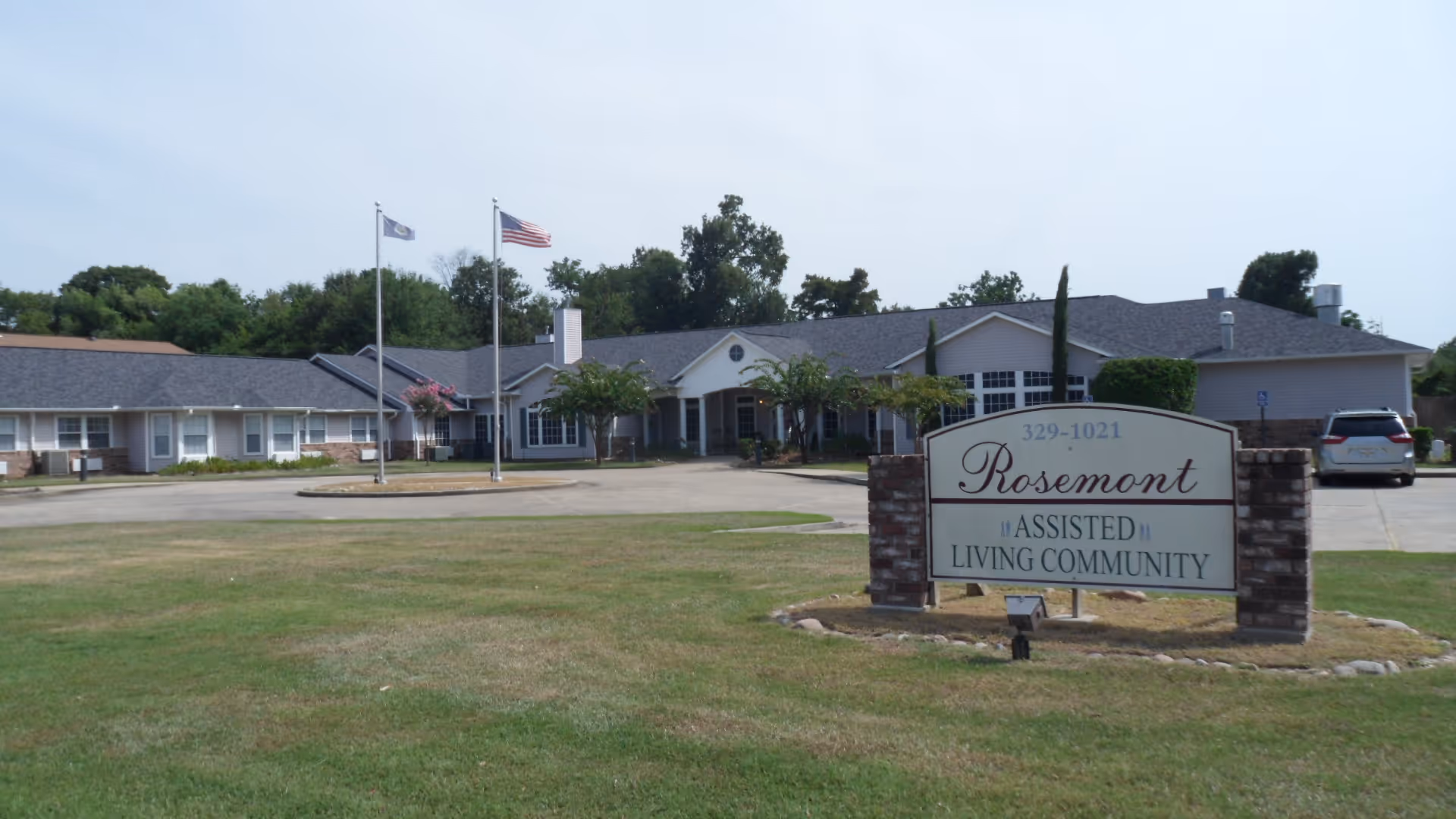 Exterior view of a single-story assisted living facility with a sign and flagpoles