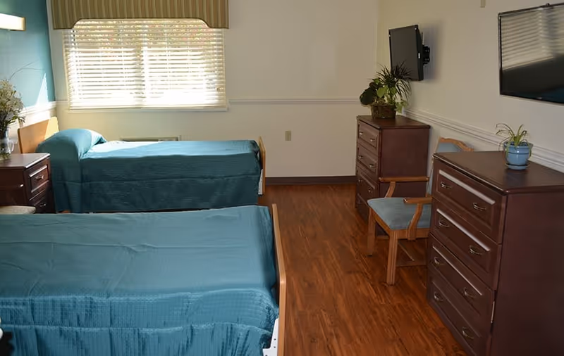 A senior living facility bedroom with two single beds covered in teal bedspreads. The room has wooden flooring, two wooden dressers, a wooden chair with a cushion, a wall-mounted TV, and a window with blinds and a valance. There are also some potted plants on the dressers.