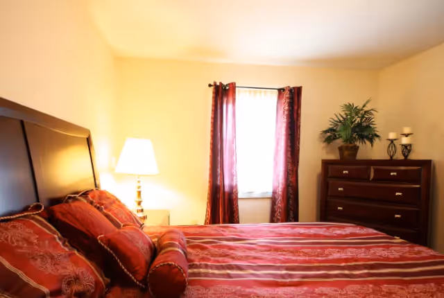 A bedroom with a large bed covered in red and gold patterned bedding. There is a bedside table with a lamp on the left side of the bed. A window with red curtains is centered on the back wall. To the right of the window is a wooden dresser with a potted plant and two candle holders on top.