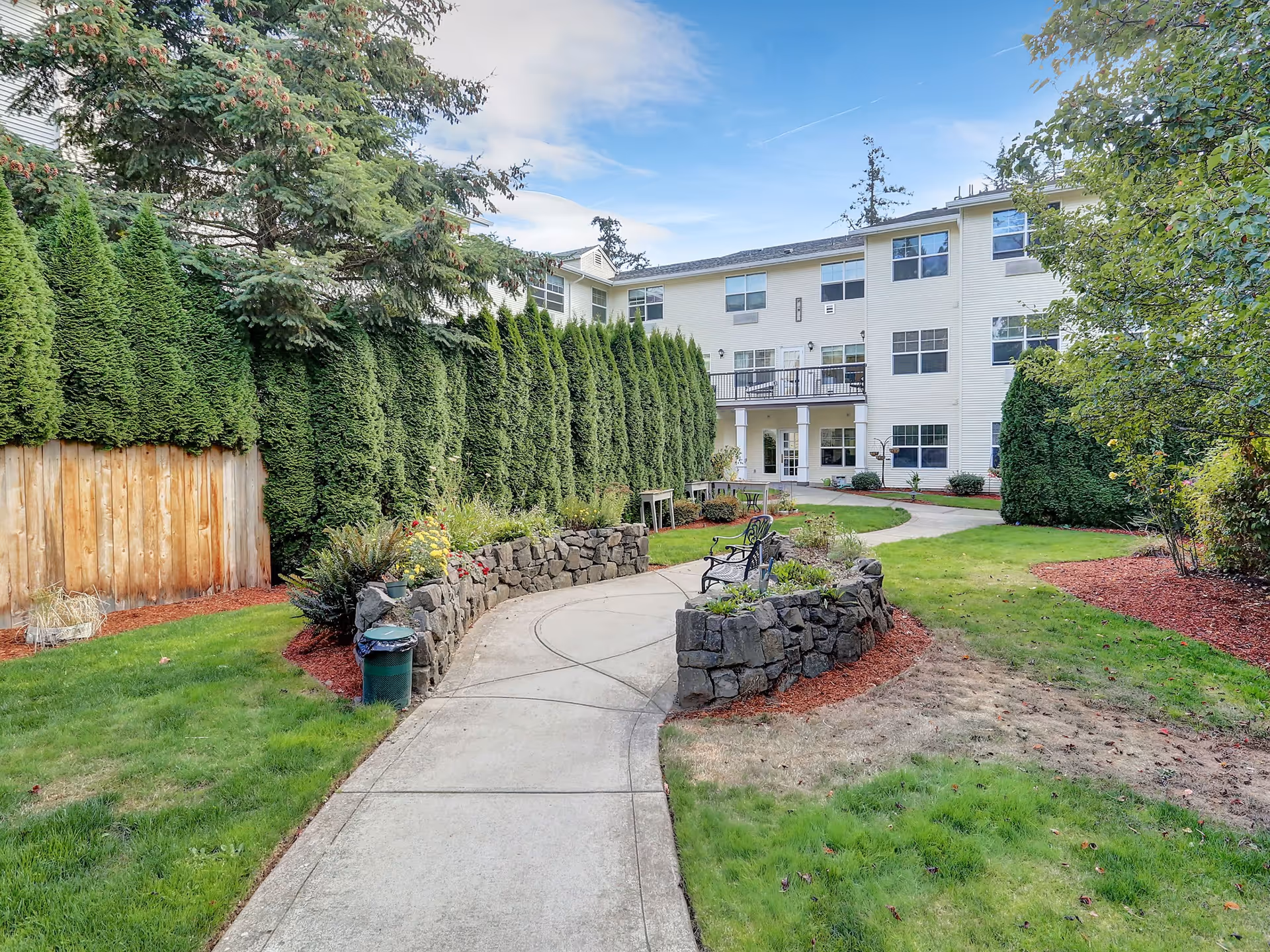 A landscaped outdoor garden area with a curved concrete walkway leading to a three-story white building. The garden features green grass, tall evergreen trees along a wooden fence, stone retaining walls with plants, and a black metal bench. The sky is partly cloudy with blue patches.