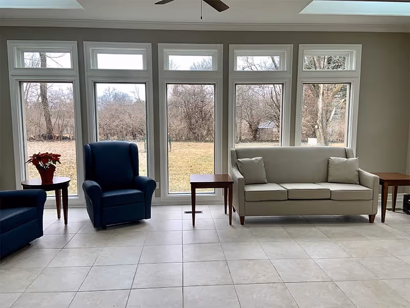 A bright room with large floor-to-ceiling windows showing an outdoor view of trees and grass. Inside, there is a beige sofa with two cushions, two blue armchairs, and three wooden side tables, one of which holds a red poinsettia plant. The floor is tiled and the ceiling has a fan.