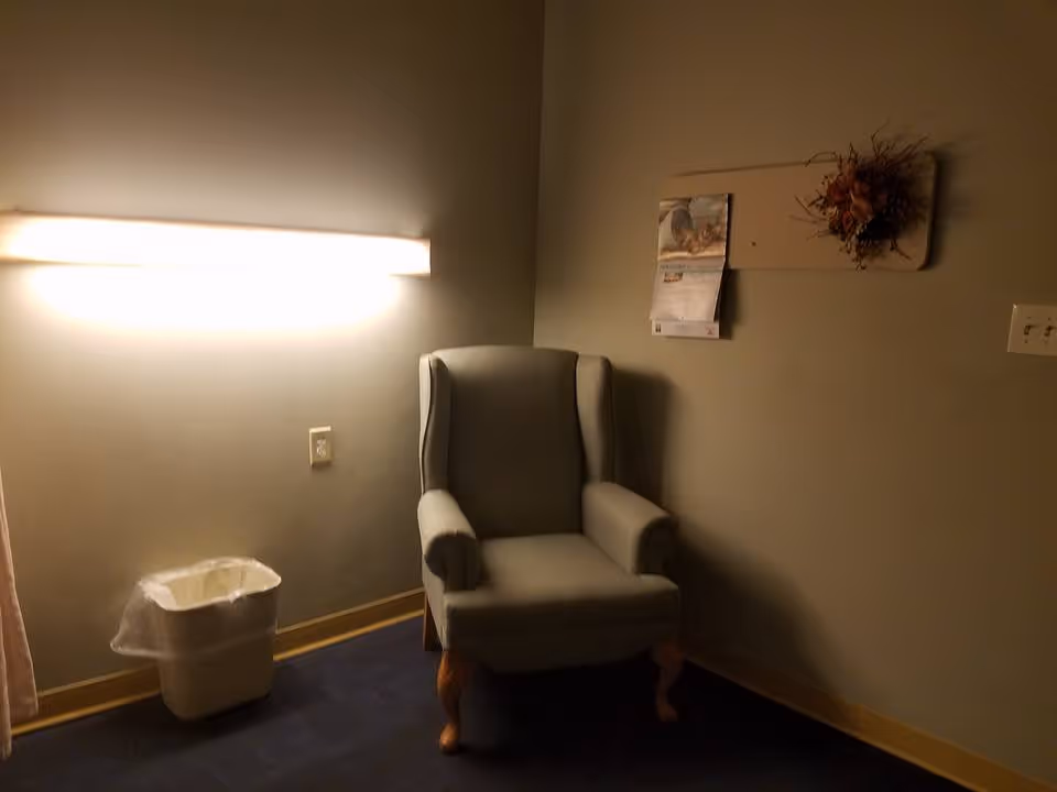 A dimly lit corner of a room with a gray upholstered armchair, a wall-mounted light fixture above, a small trash can with a plastic liner on the floor, and a wall decoration with a calendar and a floral arrangement.