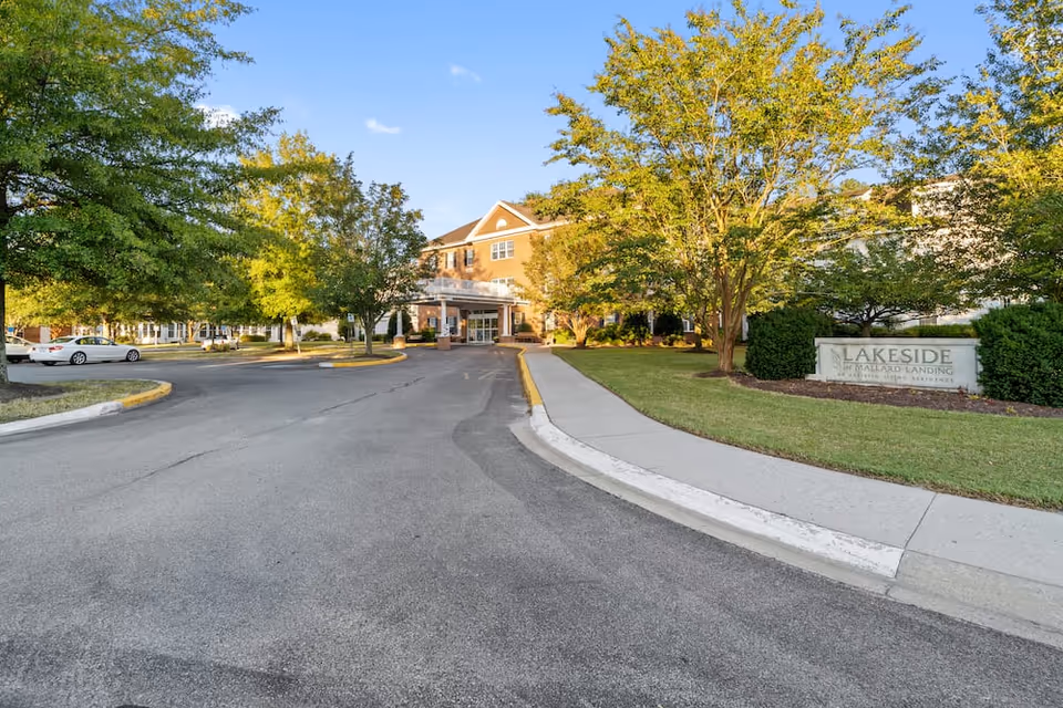 View of the entrance to Lakeside At Mallard Landing, a senior living facility, with a driveway leading up to the building. The building is surrounded by green trees and well-maintained landscaping. A stone sign with the facility's name is visible on the right side near the sidewalk.