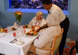 Two elderly women seated at a dining table with plates of food and drinks, while a staff member serves or assists them. There is a large aquarium with colorful fish in the background and a vase with orange flowers on the table.