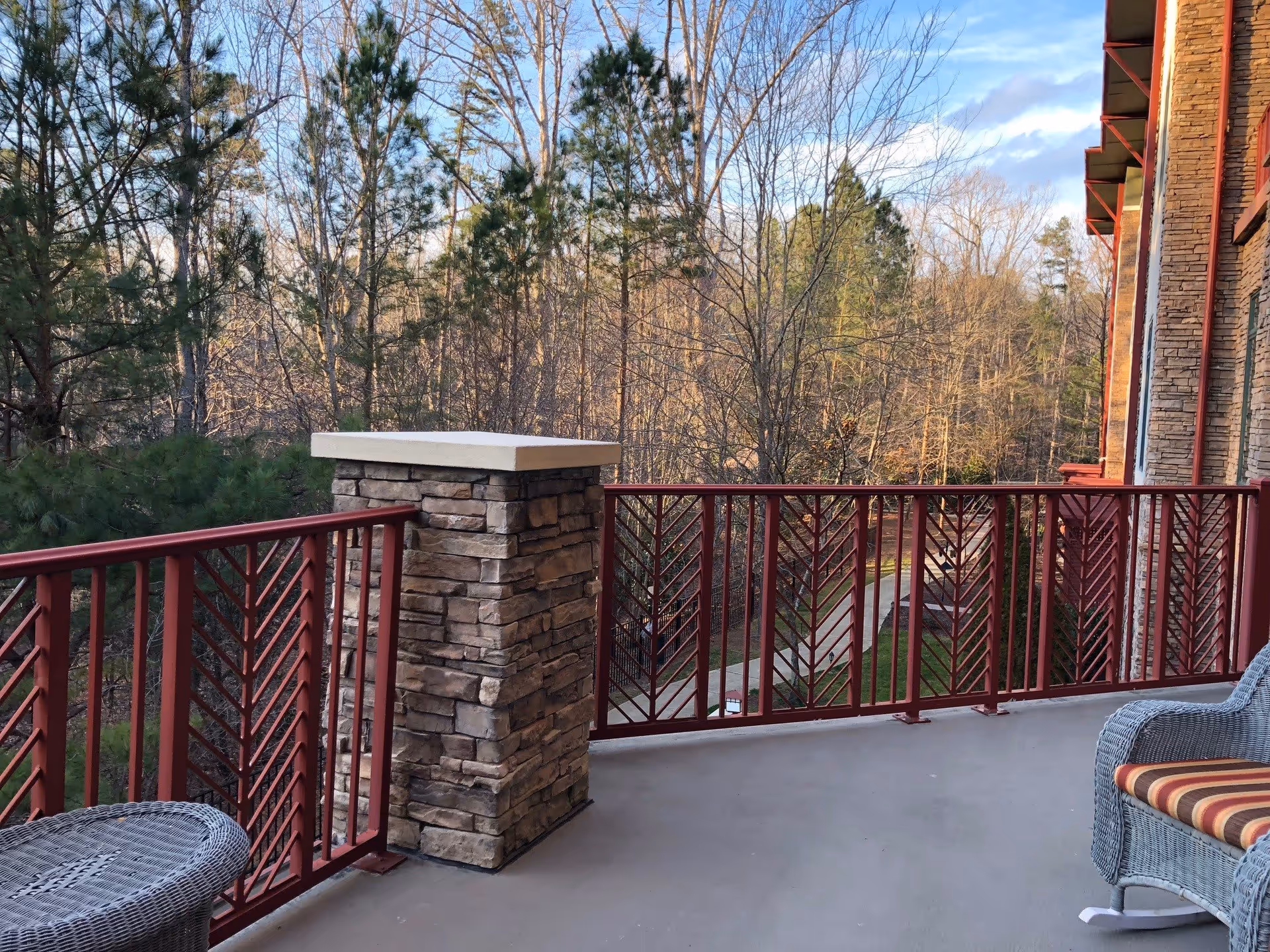A balcony with red metal railings and stone pillars overlooking a wooded area with trees and a pathway. There is a wicker chair with a striped cushion and a wicker table on the balcony. The building exterior is partially visible with stone and red trim.