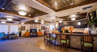 Spacious and well-lit common area featuring a kitchen with a bar counter and stools, decorated with autumn-themed floral arrangements. The room includes a seating area with armchairs and a fireplace, as well as a dining area with tables and chairs. The ceiling has decorative panels and multiple light fixtures.