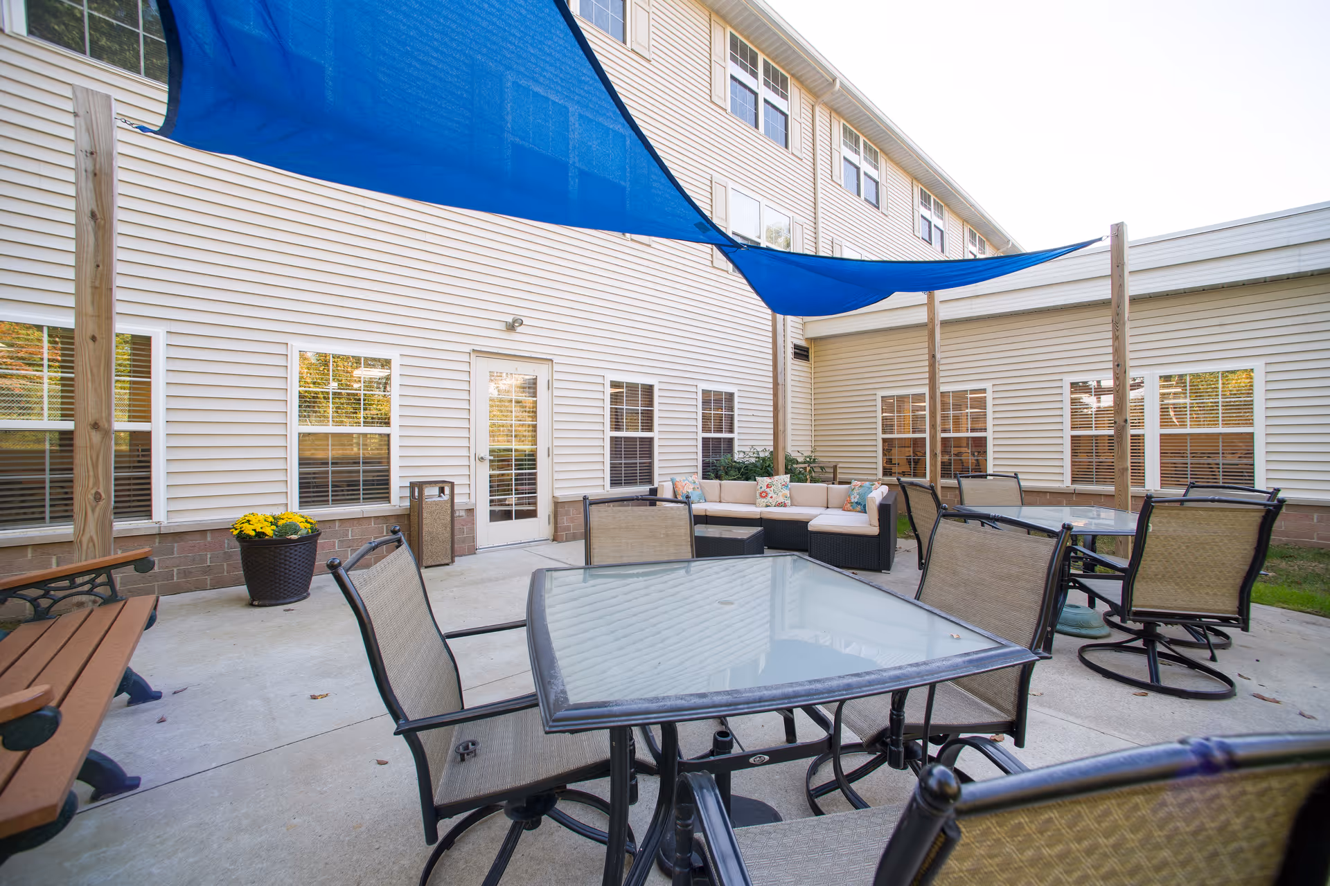 Outdoor patio area at Danbury Cuyahoga Falls with glass-top tables and chairs, a cushioned sectional sofa with decorative pillows, a wooden bench, potted plants, and blue shade sails overhead attached to wooden posts. The patio is surrounded by the building's beige siding and multiple windows.