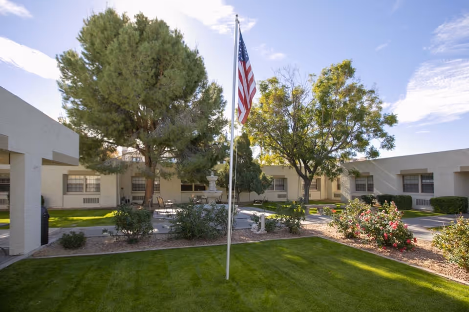 Outdoor courtyard area of Shea Post Acute Rehabilitation Center with green grass, trees, bushes, a flagpole with an American flag, and a fountain surrounded by chairs and tables. The building surrounds the courtyard with windows visible.