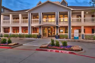 Front exterior view of a two-story senior living facility building with a covered entrance, balconies on the second floor, and landscaped flower beds in front. The sky is clear and blue.