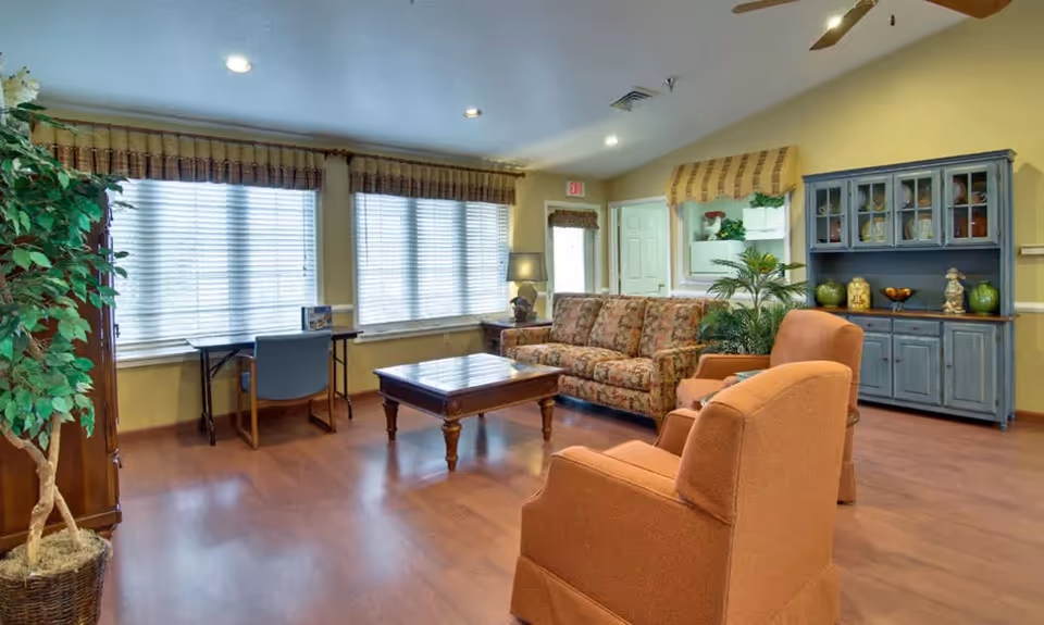 A cozy living room with a floral patterned sofa, two orange armchairs, a wooden coffee table, and a blue cabinet with glass doors displaying decorative items. There is a small desk with a chair near the windows covered with blinds and valances. The room has wooden flooring, potted plants, and warm yellow walls with recessed lighting.