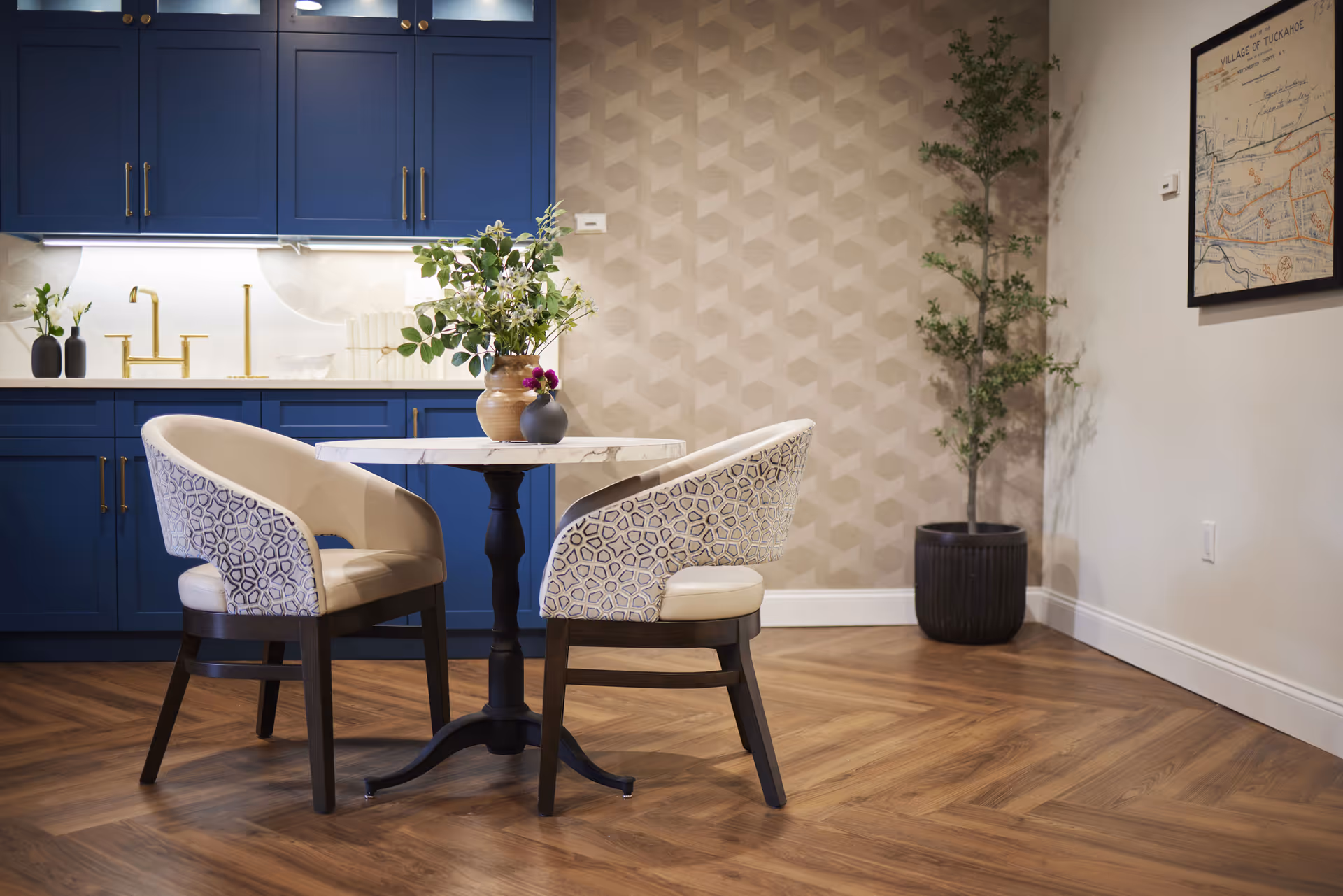 A cozy dining area with a small round table and two upholstered chairs featuring a geometric pattern on the backrests. The table has a vase with flowers on it. Behind the table is a kitchen area with blue cabinets, a white countertop, and gold-colored faucet fixtures. The floor is wooden with a herringbone pattern, and there is a tall potted plant in the corner next to a framed map on the wall.