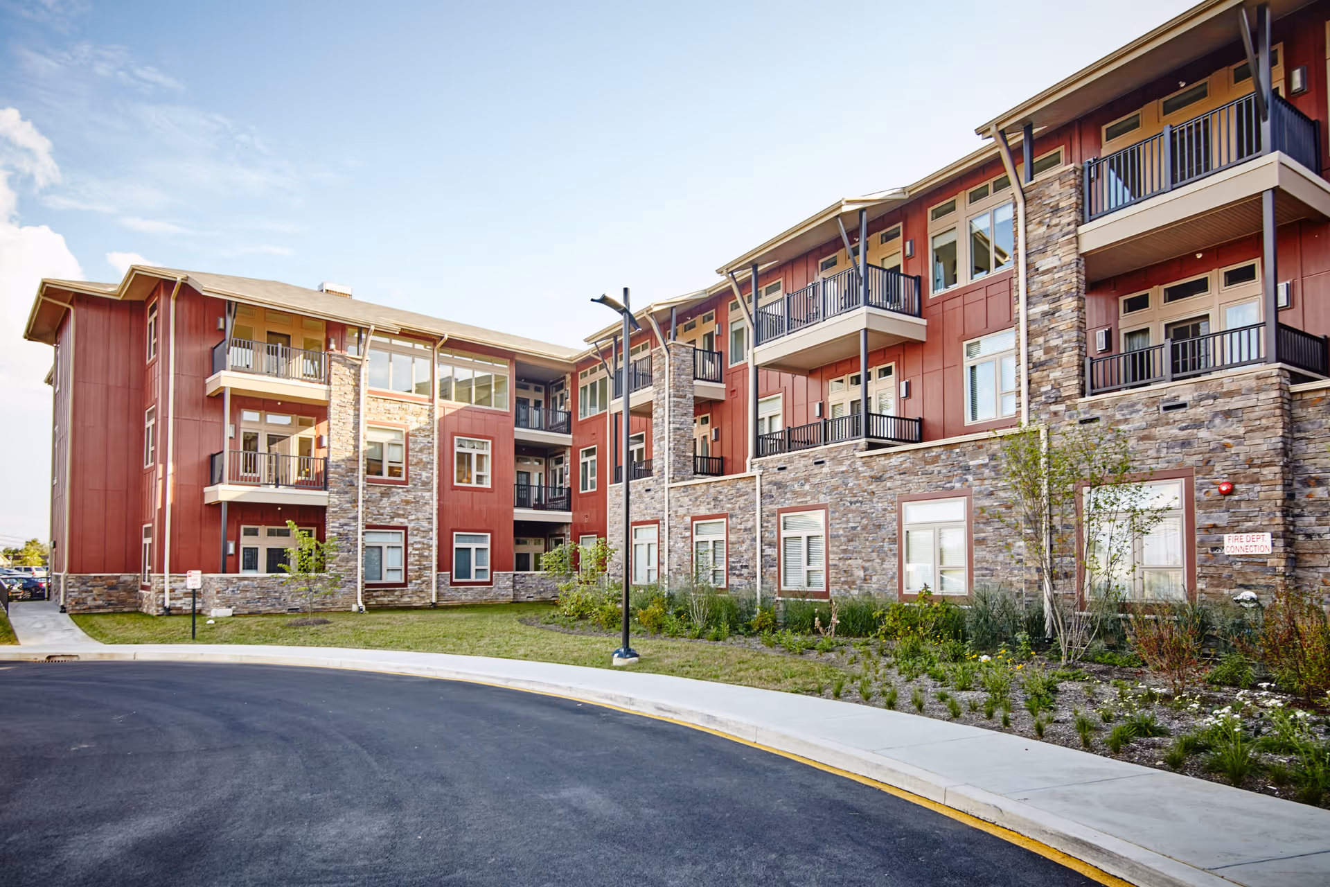 Exterior view of a modern senior living facility named The Summit, featuring a three-story building with red siding and stone accents, balconies, large windows, landscaped greenery, and a curved driveway in front.