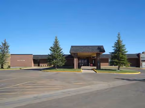 Front exterior view of Valley View Home, a single-story brick building with a covered entrance, surrounded by a paved parking area and a few evergreen trees under a clear blue sky.