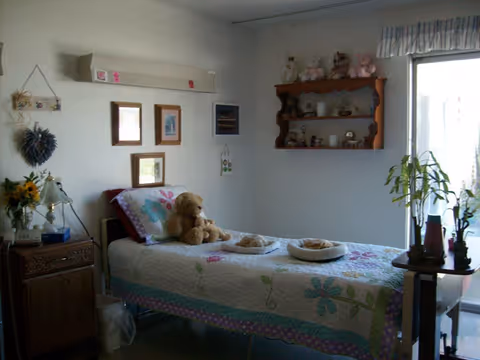 A cozy single bedroom with a floral quilted bed topped with stuffed animals, wall shelves, framed pictures, and a sliding glass door.