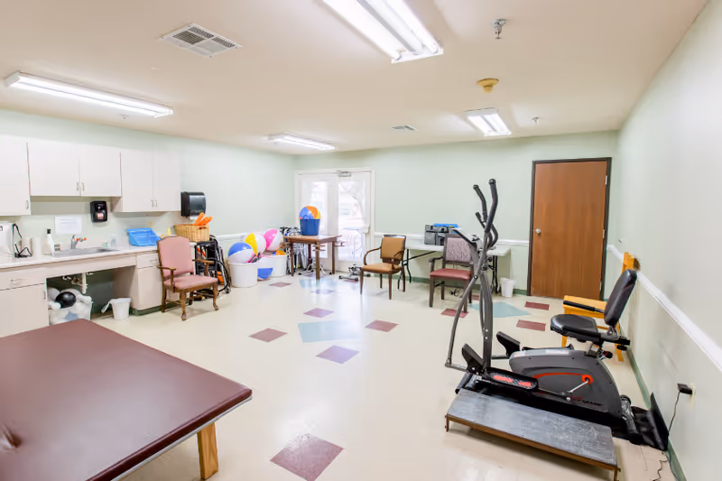 A spacious room with exercise equipment including a stationary bike and elliptical machine, several chairs, a table with beach balls and buckets, a sink with cabinets, and a door leading outside. The room has a tiled floor with a pattern of colored squares and fluorescent ceiling lights.
